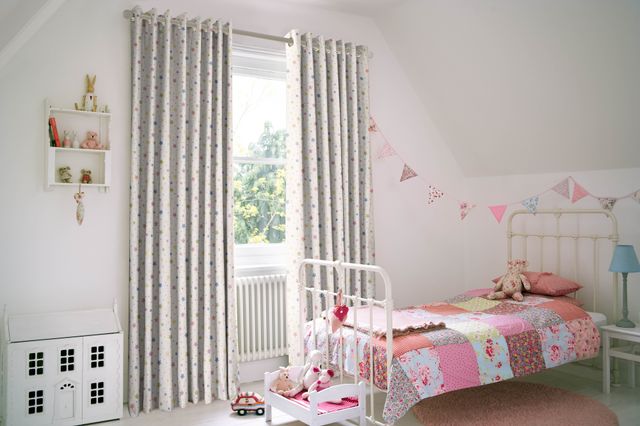 Child's bedroom in soft white with window dressed in white curtains with multi-coloured star pattern, open, white cast iron-style bed with patchwork cover in shades of pinks with flower patterns, bunting in pinks and blues, white dolls house, small white wall shelving with small animals, small pink round rug by bed. 