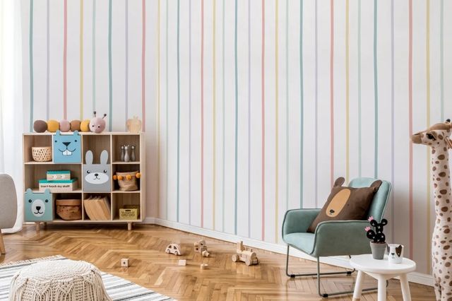 Nursery with wallpaper in white with narrow individual bands of rainbow colours, parquet wood floor with a white rug with thin bands of blue, storage unit with animal fronts to sections, filled with baskets and boxes, blue upholstered child's chair with bear cushion, wooden toys on floor, giraffe plush by small white painted wooden round side table. 