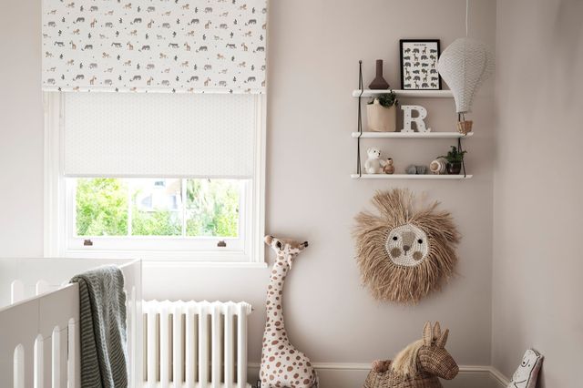Child's bedroom with natural walls and sash window dressed in two layered roller blinds, neutral with tiny yellow bursts behind a white with colourful jungle animals, wooden floor with a round faded-effect rug in white and voilet, white cot, jungle-theme animals in corner. 