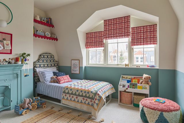 Kids bedroom in cream with lower half walls in mid dusky blue, white and blue striped bedding, red edges to shelf and pinboard and inset windows dressed in three red and white checked Roman blinds. 