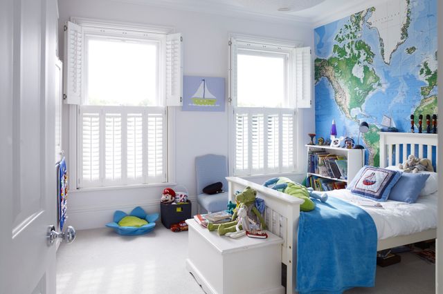 Child's bedroom in white with two adjacent windows dressed in white tier on tier shutters, top set open on both, white bed, feature wall behind map of the world, white bedding with blue throw and pillows, white toy box, white bookshelf, picture of sailboat between windows, white floor, white round paper lampshade to ceiling light. 
