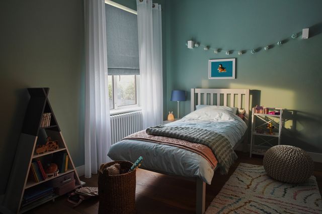 Child's bedroom in pale green-blue with window dressed in pale blue roman blind, lowered halfway and white sheer curtains, open, wooden bed with pale blue bedding with dark blue striped throw, blue bunting over bed, blue bedside lamp, triangular shelving unit with books and toys. 