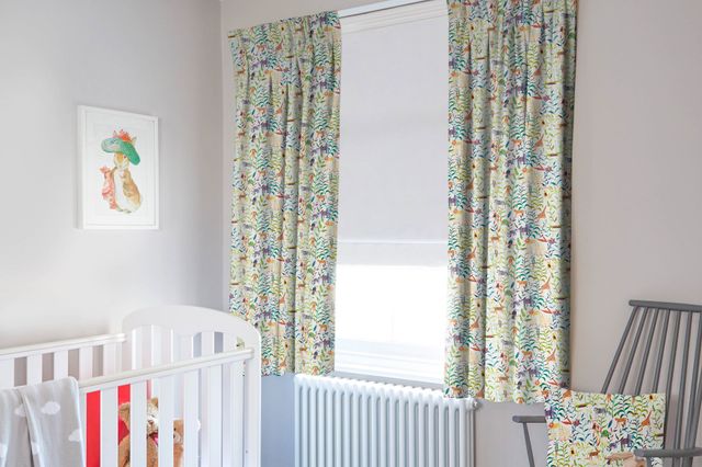 Child's bedroom in pale grey with window dressed in chalk white roller blind, lowered halfway under curtains in white with jungle animals in teal, umbre and ochre, grey wooden chair with cushion matching curtains, white cot with grey throw over. 