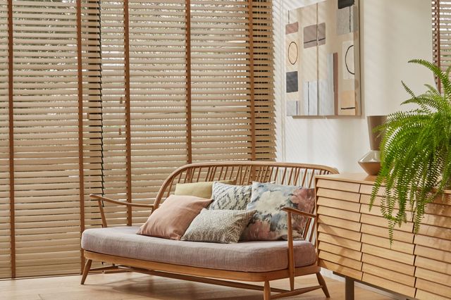Living room in cream with windows and doors dressed in faux wood mid colour venetian blinds, fully lowered and tilted, low wooden sofa with cushions in pale peach and watercolour-effect patterns, wooden side unit with large fern, wooden floor with rug in cream with black dashes, light rays on walls. 