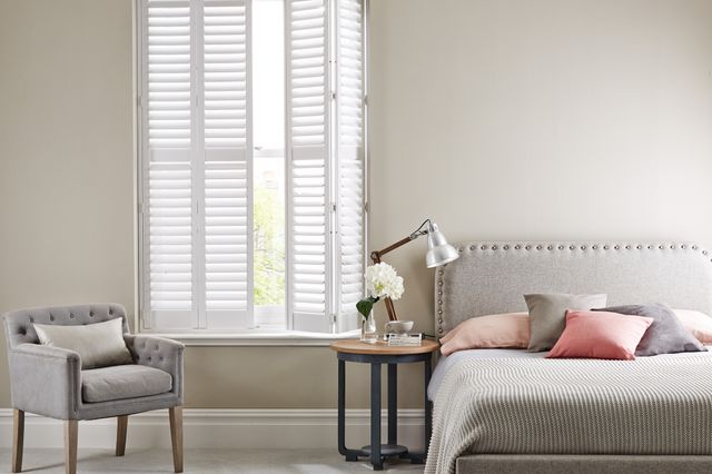 Neutral bedroom with tall window dressed in white shutters, right hand partially open, pale mink upholstered bedhead with pinstriped mink and white bedding, cushions in mink and dusky peach, mink upholstered chair, round bedside table with anglepoise lamp and two white blooms in small vase. 