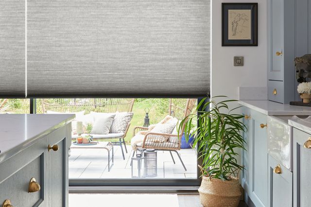 Pale greige kitchen with windows and doors dressed in silk charcoal mid grey blinds, lowered halfway revealing patio and chairs, dark wood floor, mid grey base and wall units on right, island units on left, jute rug by windows and tall plant in jute basket in corner. 