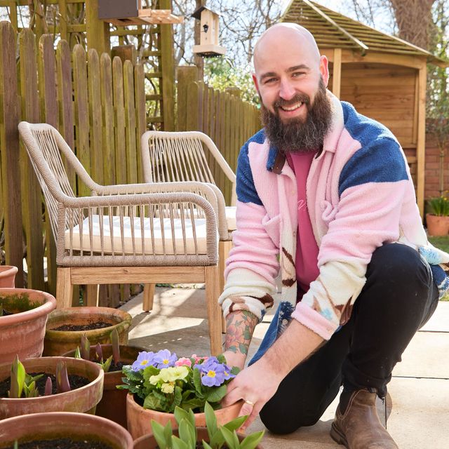 Influencer Luke Newnes, wearing a pink and blue fleece in his garden planting purple and yellow flowers in a pot with multiple pots and shoots around on a sandstone-style patio next to two raffia chairs with cream cushions.