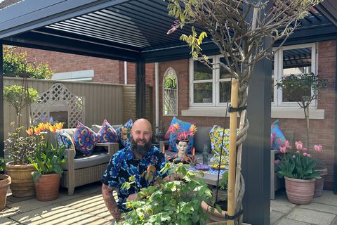 Exterior of a new build property, on a laid sandstone-style patio, a man in a hawian shirt kneels beside a tall tree in a large terracotta pot, with a smaller shrub in a terracotta pot, in front of a grey fresstanding pergola, with pots of flowers and trees and a grey sofa with colourful cushions.