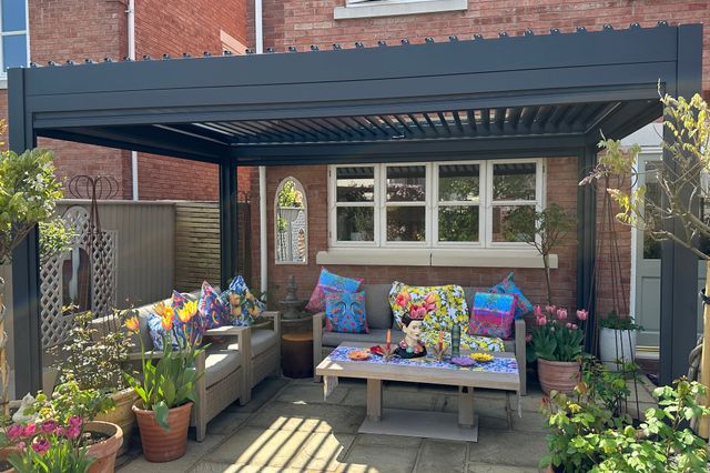 Exterior of new build house with sandstone-style patio under a grey freestanding pergola, two wooden sofas with grey cushions, and colourful cushions, low table with colourful decorations, multiple pots with flowers, shrubs and trees.