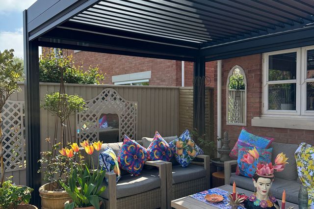Exterior of new build house with sandstone-style patio under a grey freestanding pergola, two wooden sofas with grey cushions, and colourful cushions, low table with colourful decorations, multiple pots with flowers, shrubs and trees.