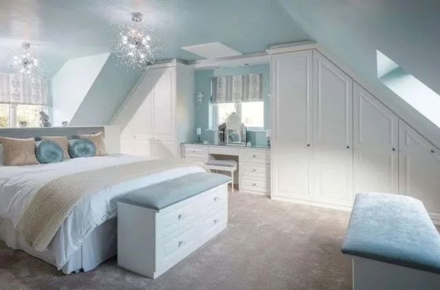Pale blue bedroom with opposing sloping walls, white fitted wardrobes and dressing table, bed with pale blue bedding, white freestanding drawer unit with pale blue padded seat, matching unit opposite under skylight, ecru carpet.