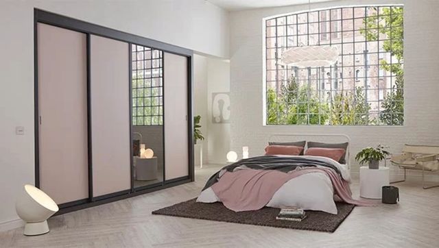Cream bedroom with wall to wall fitted units in dark grey with three taupe sliding doors closed, and one mirror door, bed with white bedding and grey and pink throws and cushions, huge leaded window behind bed, with no dressing, multiple lamps in corners and around bed, pale parquet flooring, with dark brown rug.
