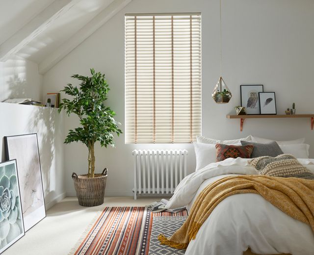 White bedroom with tall narrow window dressed in pale wood venentian blind, closed and tilted open, bed with white bedding with ochre throw, small ficus in raffia basket, cream carpet, rug in shades of saffron, umbre and ochre in linear pattern.