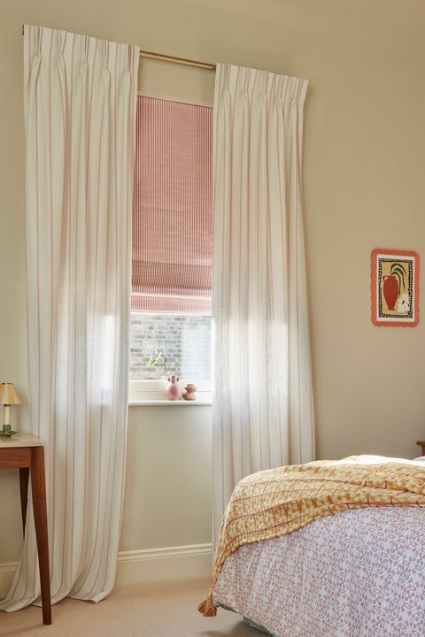 Bedroom with cream walls, bed with dusky pink headboard, bedding in white and pink patterned cover with cushions with stripes and checks, dressing table and shaped mirror and window dressed with pale red and white striped Roman blind with curtains in white with pale red pinstripe on a rod mounted higher above the window.