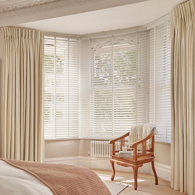 A bright and elegant bedroom corner featuring large bay windows with white wooden blinds, framed by floor-length cream curtains. A classic wooden chair with a woven seat sits by the window, draped with a soft white throw, and a small stack of books and a cup rest on the seat. The space is softly lit by natural daylight filtering through the blinds, highlighting the neutral-toned décor and cosy atmosphere.