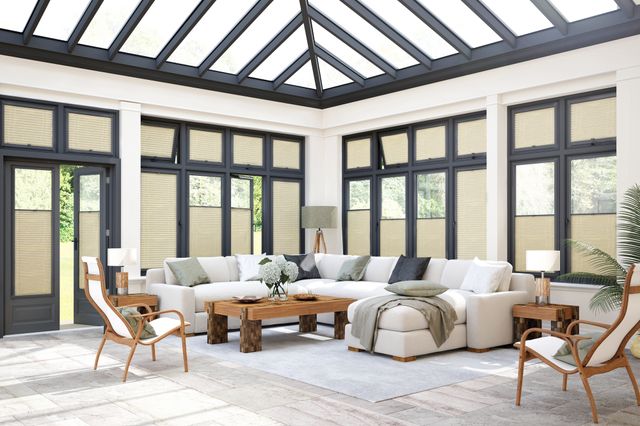 Cream room with ivory pleated bllinds in anthracite perfect fit frames in conservatory styled windows to sides and roof, at varying heights, cream sofa with cream and anthracite cushions, wooden coffee table and side table, wood and cream chairs.