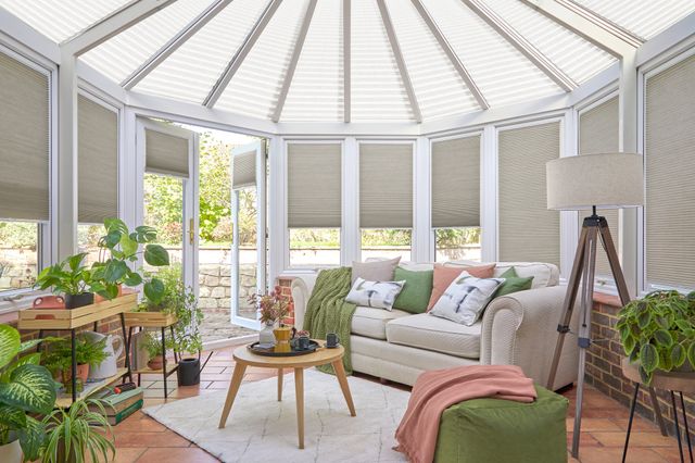 White conservatory with pale terracotta tiled floor, white sofa with cushions in green, white and salmon, green poufee with salmon throw, multiple plants and stone coloured duette blinds in silk finish to conservatory windows and white pleated blinds to roof, doors open.
