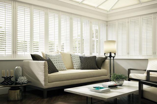 Conservatory with white shutters on side windows, closed with louvres open, white pleated blinds to roof, cream sofa with cushions in black and white polka dots and white with black asterisk, coffee table with white top, standard lamp with white shade.