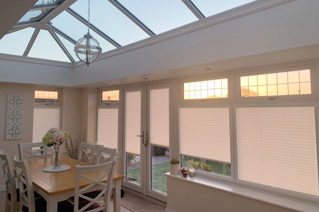 Kitchen-extension conservatory with white blinds on side windows at varying heights, pale copper pleated blinds to roof, square dining table and four chairs, two-seater sofa, cream rug.