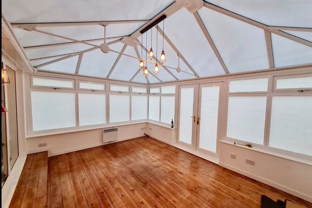 Large rectangular conservatory with soft white blinds to roof and sides, polished wooden floor, empty of furniture, industrial-chic style lights, on.