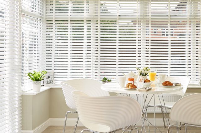 Close in on white faux wood blinds in a conservatory, fully lowered and tilted, modern white chairs and round table with white crockery, croissants, orange juice and tea..