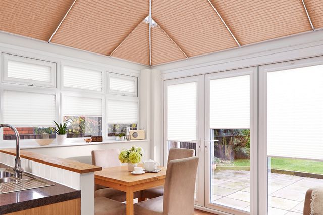 Kitchen-extension conservatory with white blinds on side windows at varying heights, pale copper pleated blinds to roof, square dining table and four chairs, two-seater sofa, cream rug.
