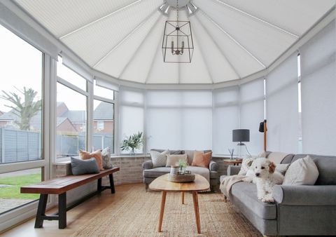 Conservatory with white blinds on side windows, three fully raised and the rest closed, cream pleated blinds to roof, mid grey sofa with cushions in cream, matching large chair, jute rug underneath, wooden bench, cute white dog on the sofa.