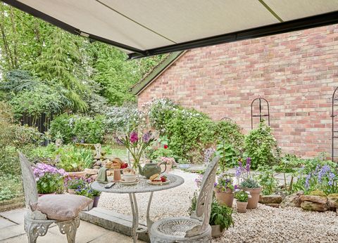 Cottage-style garden showing edge of traditional house with lush greenery and flowers, taken from a patio behind white wrought iron-style table and chairs under a pergola awning in beige-grey.