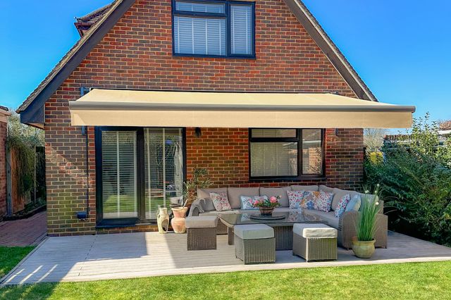 Exterior modern brown-red brick house with grey paved patio under a pale umber awning, extended, with grey-brown rattan-style L-shaped sofas and glass-topped table, cream cushions with pale blue and pink accent cushions, pink blooms on table, lawn to foreground.