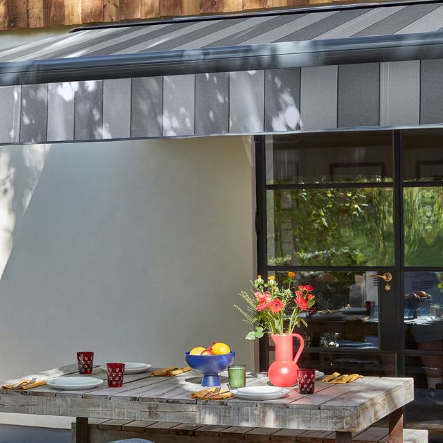 Close up of a wooden table and two benches on a patio in the sun under a grey striped awning, an orange vase with red poppies, a blue fruit bowl, forest green throw on bench with a dark cushion.