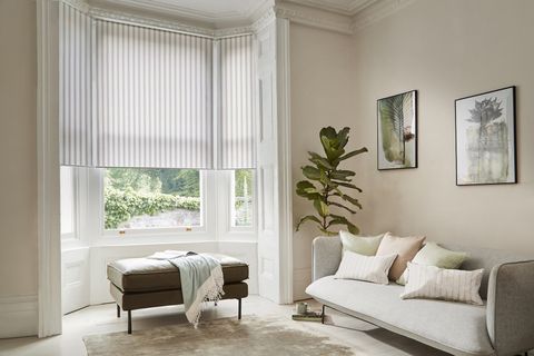 Sandstone living room with white panelling to side of bay window, dressed in three white roller blinds, lowered three quarters, brown footstool in recess with pastel green throw with white fringe, pale grey sofa with white, blush pink and white linear pattern in soft pink and blue, large plant in corner, watercolours on wall.