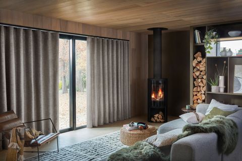 Cozy living room with grey curtains, wood-burning stove, modern sofa, and shelves stacked with firewood and decor.