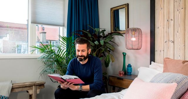 Oliver Heath sat reading a book in abedroom that has white roller blinds paired with blue curtains on a white rectangular window